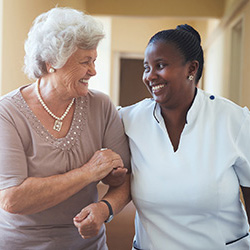 an elderly woman smiling and walking arm in arm with a caregiver
